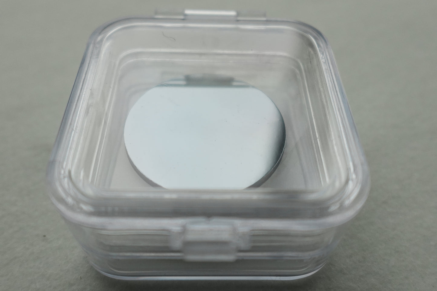 Clear plastic sample holder box containing a round, flat metallic wafer on a gray surface