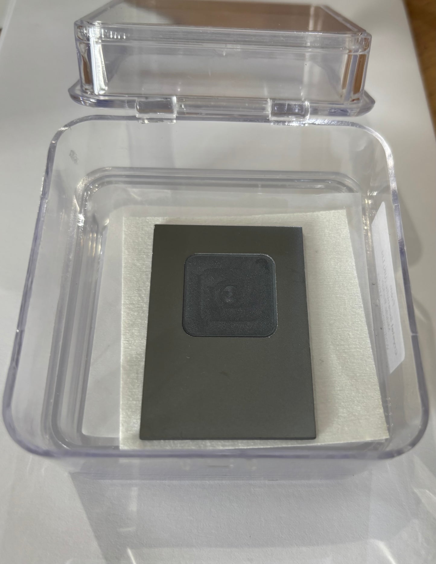 Small dark rectangular metal object with embossed symbol inside clear plastic display box